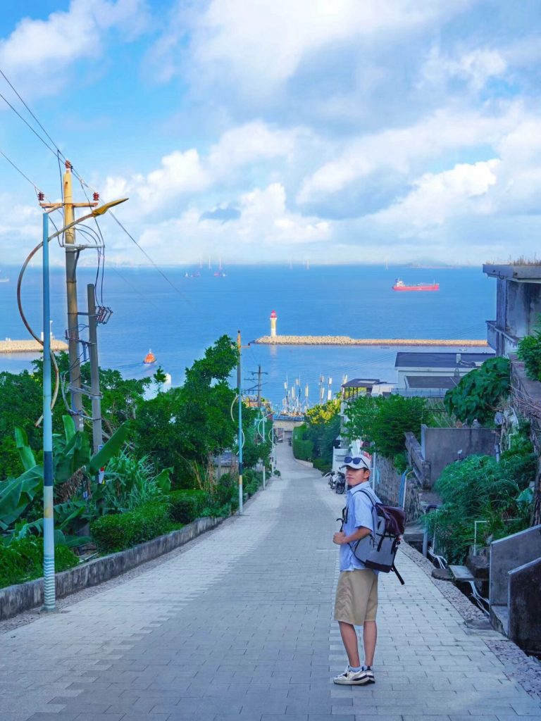 Man standing on a path overlooking the sea with boats and a lighthouse