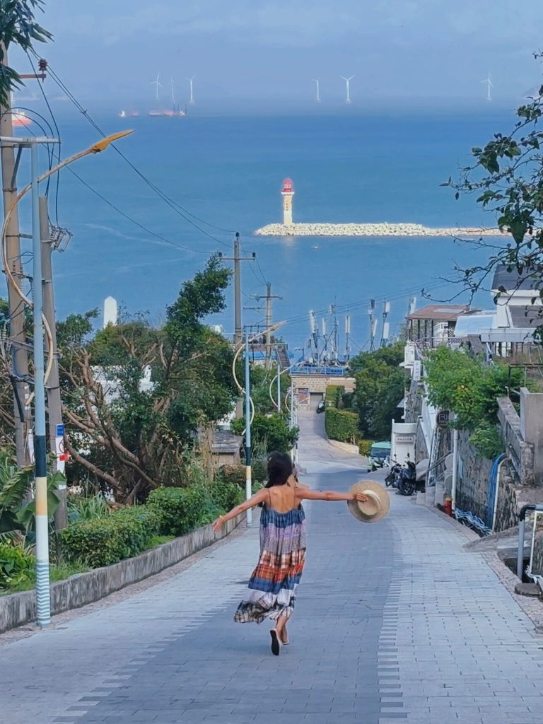 Person dancing joyfully on a path with ocean and windmills in the distance