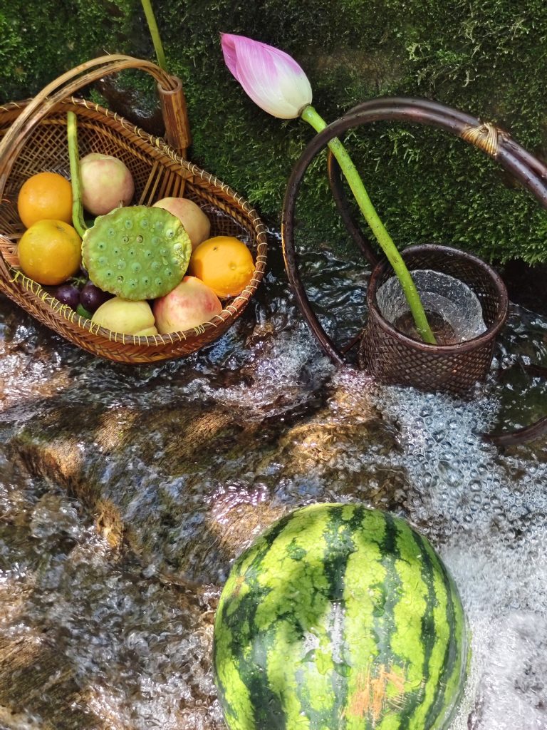 Basket of fruits including oranges, peaches, and lotus pods by a stream